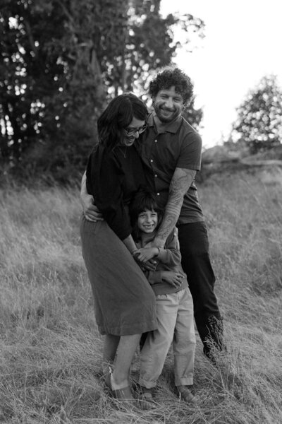Candid outdoor portrait of a couple hugging their child, captured in black and white with tall grass in the background.