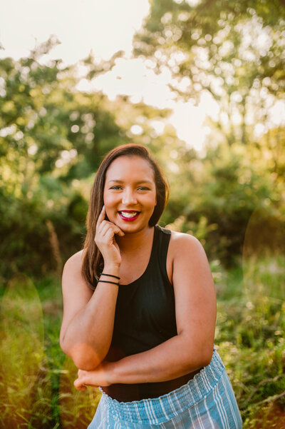 Smiling woman standing outdoors in a sunlit field, wearing a black top and light blue jeans. The warm, golden light highlights her joyful expression, perfect for personal branding photography.