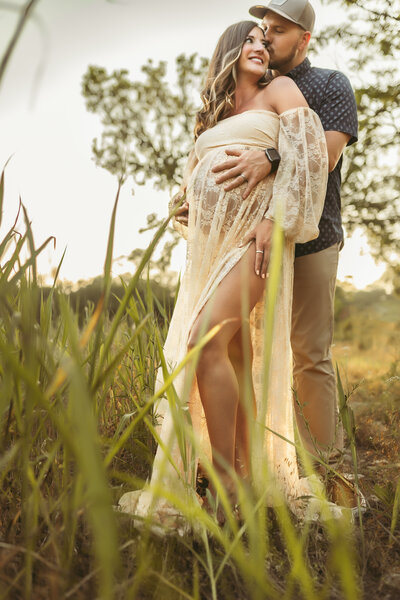 Man in blue button up shirt and brown pants with a baseball hat hugs and kisses his wife in a cream lace gown in an outdoor Shawnee location
