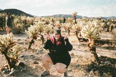 A person crouched down holding up peace signs in a field of cacti