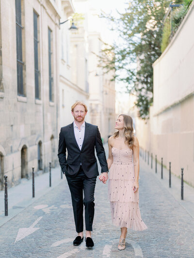 a man and woman holding hands and walking towards the camera on a side street in paris