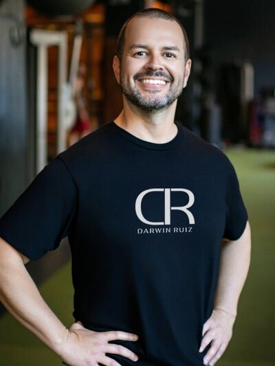 Darwin Ruiz wearing a black T-shirt featuring the mirrored DR monogram and his full name “Darwin Ruiz” in a clean white typeface, standing confidently in his training facility.