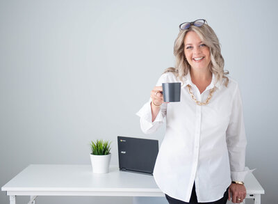 a female realtor in a white blouse holding a coffee cup as she leans on her desk.  Captured in studio by Ottawa Branding Photographer JEMMAN Photography COMMERCIAL