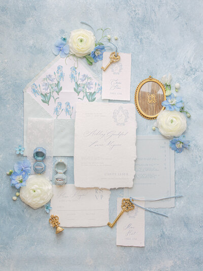 Flay lay of a white and blue themed wedding invitation suite and wedding rings surrounded by blue and white flowers and gold keys on a blue and white background