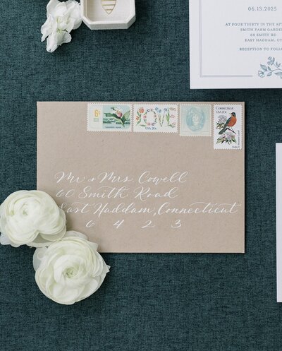 Kraft paper envelope with white ink calligraphy and vintage postage stamps on dark styling mat for Connecticut wedding at Smith Farm Gardens.