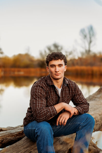 High school senior boy sits on leaning tree trunk over a lake on a fall day wearing a plaid button up and jeans