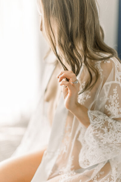 A photo of a woman sitting in a Dallas photography studio wearing a delicate lace robe holding a piece of her hair while she looks out of the window for her boudoir session.