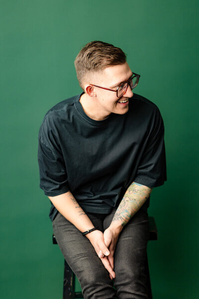 Man sits on stool in front of a green background leans over and laughs.