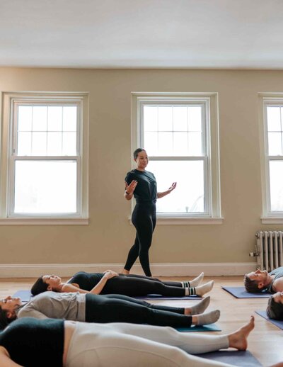 A breathwork instructor guides a group of participants lying down, focusing on deep breathing techniques in a sunlit studio.