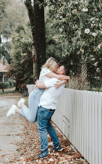 A couple wearing white t-shirts and jeans is embracing along a white picket fence. There are white flowers in trees behind them.