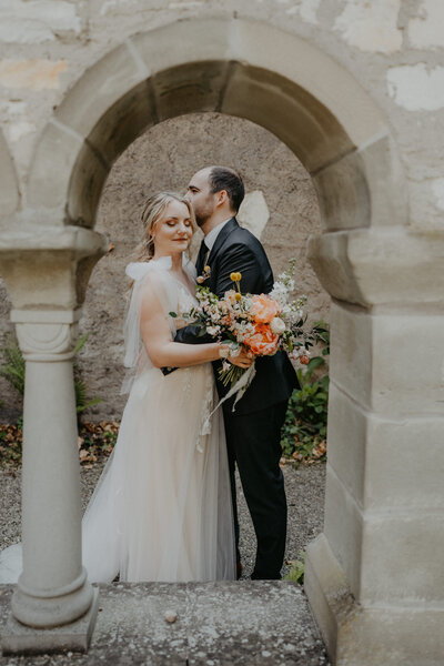 Ein Paar in traumhaftem Brautkleid steht unter einem alten Steinbogen – floraler Akzent durch stilvollen Brautstrauß.