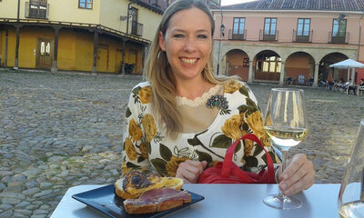 Maria Cristina smiling and sitting at table in a plaza with a tapa and a glass of white wine.