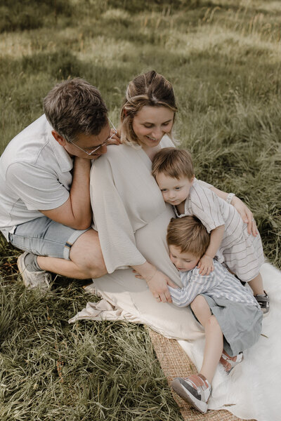 Een koppel waarvan de mama zwanger is en hun 2 zoontjes zitten knuffelend in het gras tijdens een zwangerschaps fotoshoot in de natuur te Mechelen.