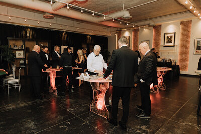 Guests in formalwear gather around glowing casino tables during a black-tie event at SoHo63, a Chandler wedding and event venue. The room features exposed brick, dramatic lighting, and a festive atmosphere perfect for upscale celebrations and private parties.