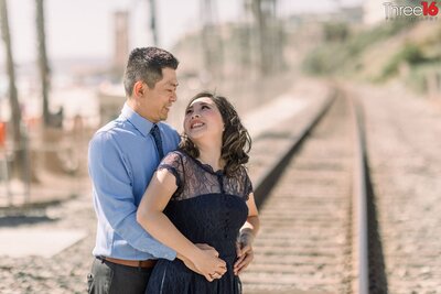 Bride to be looks back and up at her fiance with smiles while standing on a train track in San Clemente