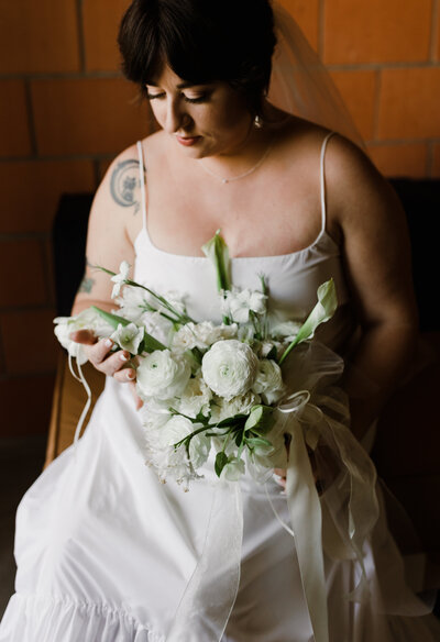 Bride wearing simple white dress looking down at bouquet of white florals at Carpenter hotel wedding