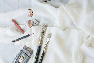 two paint brushes, pencils, pencil sharpener with shavings, and artists tools laid out on cream tablecloth