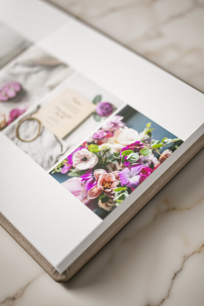 A photo album with a luxurious leather cover rests on a marble surface. It is filled with vibrant images, including a close-up of colorful flowers in shades of pink, purple, and green, and a picture of a small note card and a vintage key.