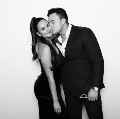 A man in a suit kisses a woman on the cheek as she smiles at the camera. Both are dressed formally, standing in front of a plain white backdrop.