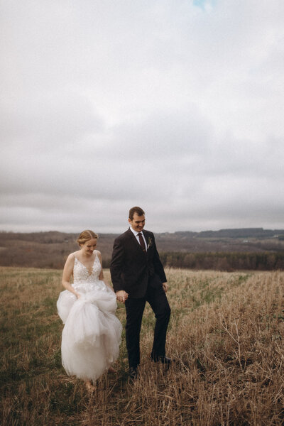Couple holds hands while walking through a cut hay field on their wedding day near Collingwood, Ontario. 