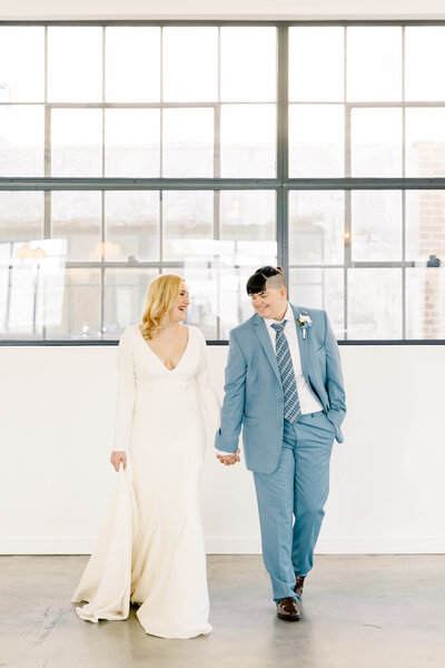 Bride and bride hold hands walking and laughing at one another in their reception room surrounded by windows and natural light by Kansas City Wedding Photographer Sarah Riner Photography