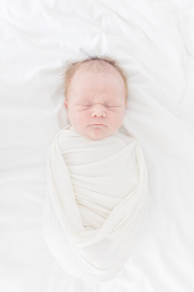 Newborn baby swaddled in a soft, white blanket, peacefully sleeping. The baby is surrounded by a serene, all-white setting, creating a calm and tranquil atmosphere.
