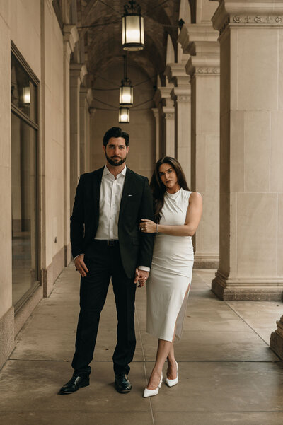 Man in a black suit and woman in a white dress stand in an elegant corridor with arches, looking confidently at the camera. Soft, warm lighting.