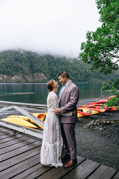 A bride & groom laugh as they look over kayaks on the shore of Lake Crescent in Olympic National Park.