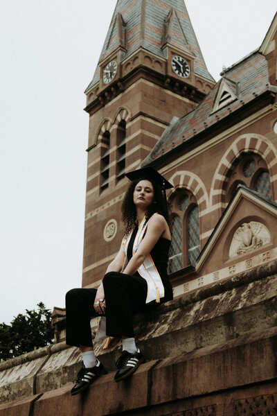 Photo of a woman sitting on the ledge of a building with her feet dangling with more college buildings in the background behind her. She is wearing her graduation cap and sash