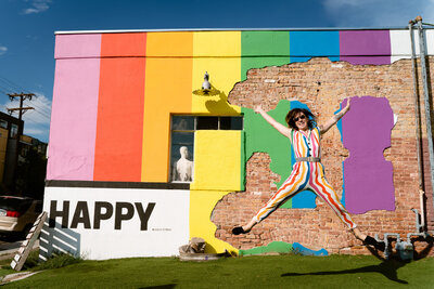 A person jumping up with their arms and legs spread out in front of a rainbow painted wall with the word happy on it.