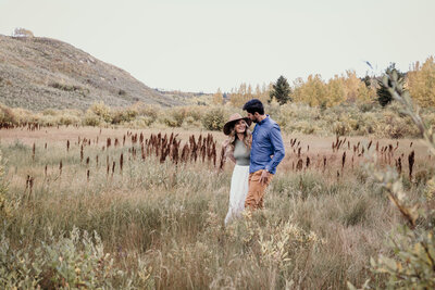 A couple walks through a field in Big Hill Springs Park during their engagement session. He's got his arm around her shoulder and she's holding that same hand while looking up at him and smiling