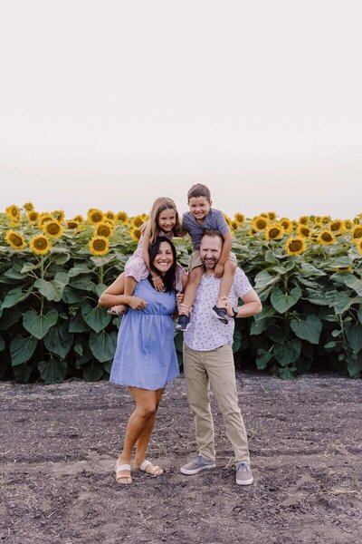 Family standing in a sunflower field, parents holding children on their shoulders while smiling at the camera.