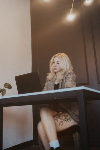 A blonde haired woman sitting at a desk typing on her laptop, wearing a plaid suit coat and high-top converse.