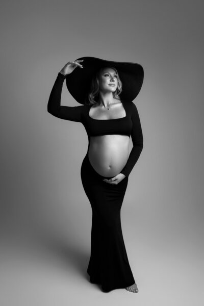 Austin maternity photographer captures dramatic black and white studio portrait of pregnant woman in elegant black dress and wide-brimmed hat