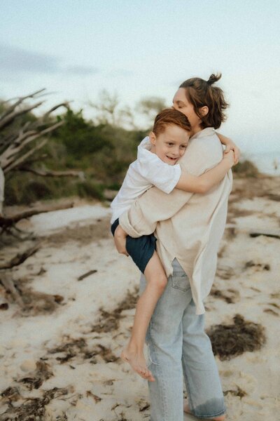 A woman standing barefoot on a sandy beach is holding a smiling child in her arms. Driftwood and greenery are visible in the background.