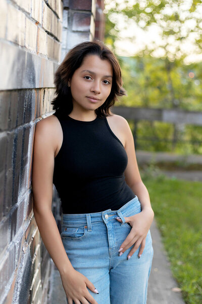 Senior girl in black sleeveless top and blue jeans leaning against graffiti wall outside during sunset