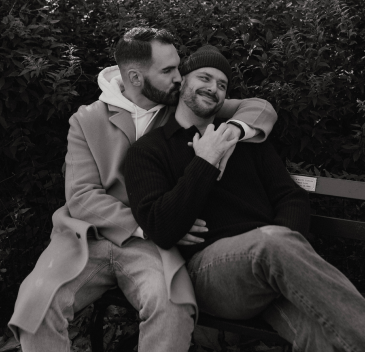 A male couple kissing on the cheek sitting on a bench in a park