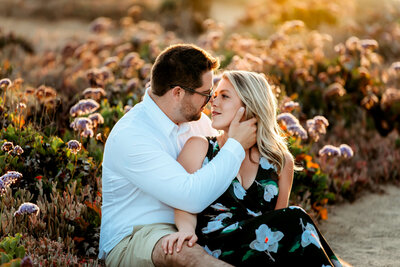 A husband and wife couple look lovingly at each while sitting in a field at Del Mar in San Diego, California. Photograph by Kim Belverud, a San Diego family photographer.