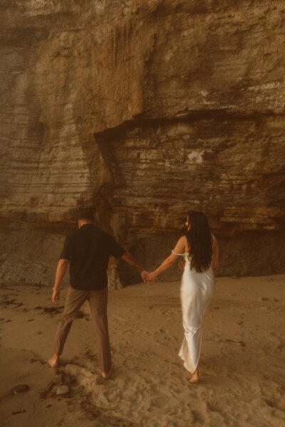 a man and woman walking away on the beach towards a cliff