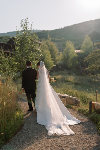 Bride and groom walk away hand-in-hand through a mountain path at sunset, the bride’s veil flowing behind her — timeless and romantic wedding photography by Kelly Lugo Photo.