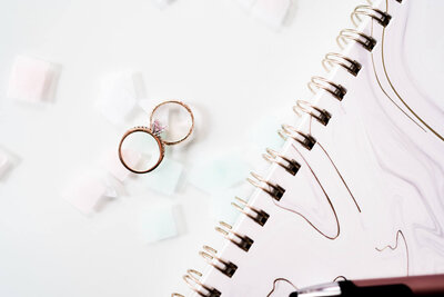 aerial view of wedding rings on top of light pink confetti next to a marble spiral notebook