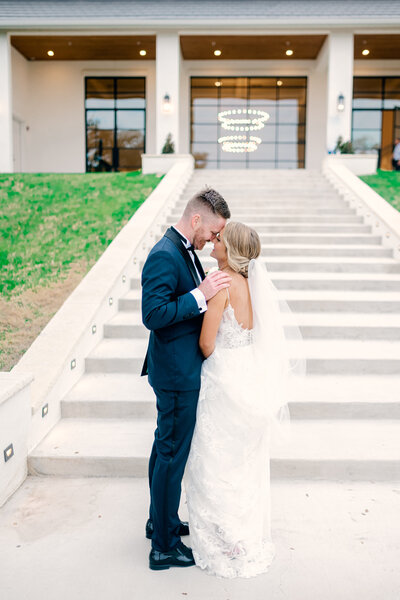 Bride and groom standing close together with their foreheads touching in front of a large outdoor staircase