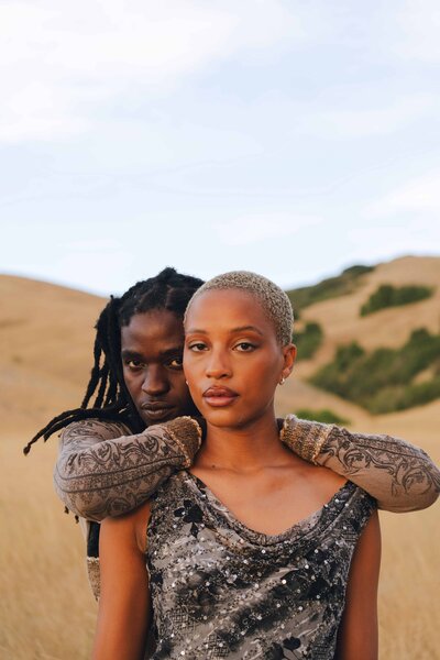 Two individuals in an intimate embrace, dressed in textured clothing, set against a serene, golden landscape with rolling hills.