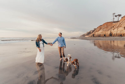 A pregnant woman holds her husband's hand while they walk on the beach in San Diego with their two dogs.