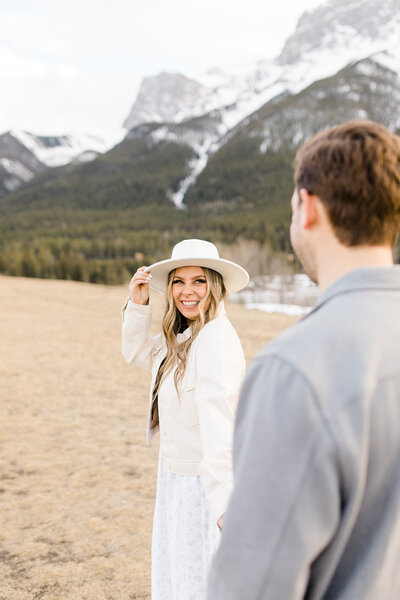 Woman in white jacket and white hat looking back and smiling at her fiance in the Rocky Mountains.
