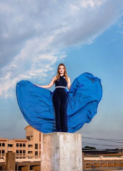 Richmond high school senior girl wearing blue sequin jumpsuit with skirt overlay stands on top of a pillar on a rooftop during her senior portrait session.