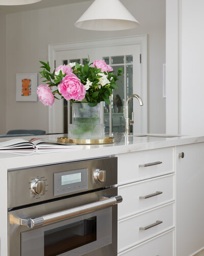 A kitchen island with a marble countertop and white cabinetry with a built-in stainless steel oven and sink. A vase of pink flowers and a cookbook sit atop the counter