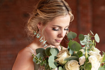 A blonde bride looks down at her bouquet of large, white roses and eucalyptus.