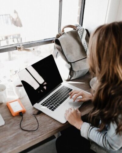 A woman with long brown hair types on a laptop at a wooden counter by a window, with a backpack, coffee cup, and external hard drive nearby—capturing a calm, productive workspace vibe.