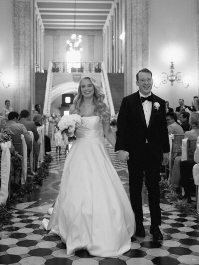 Black and white photo of a joyful bride and groom walking down the aisle in an elegant indoor venue, hand in hand, surrounded by seated guests.